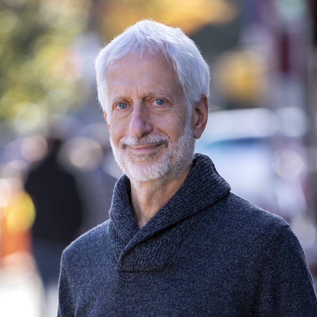 Portrait of a smiling older man with white hair and a short beard, wearing a dark knit sweater, outdoors on a city street.