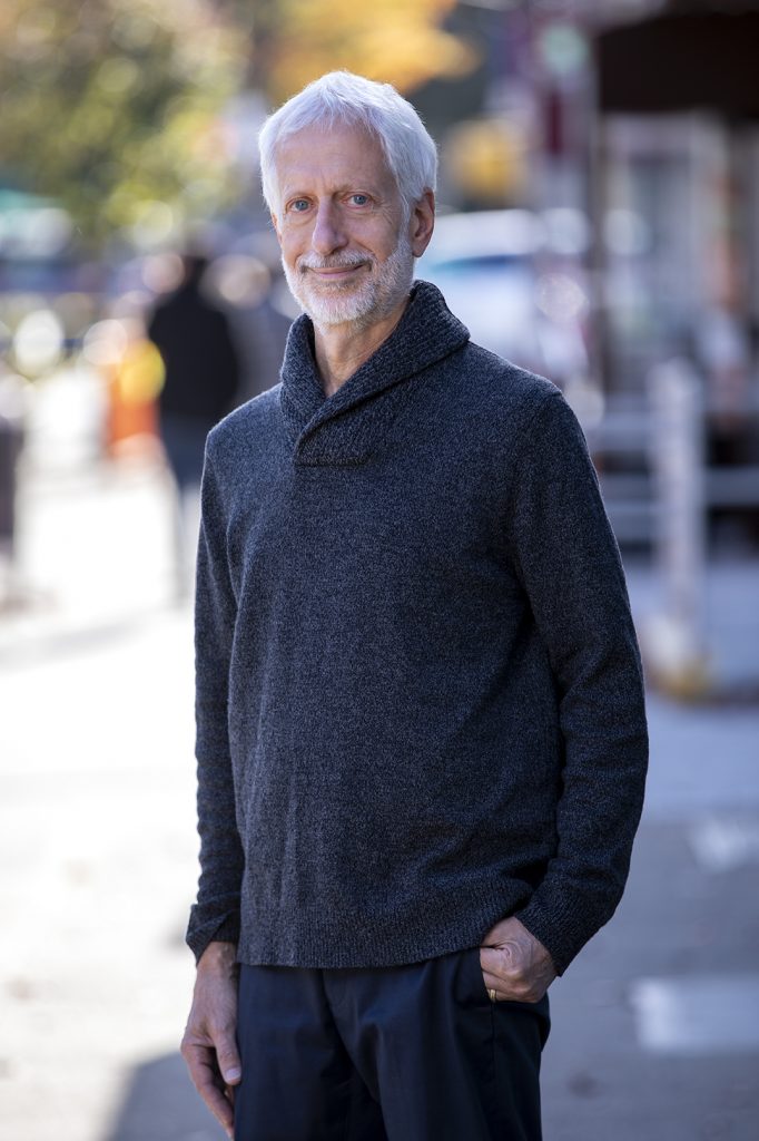 Older man with white hair and beard standing outdoors on a city street, wearing a dark gray sweater and looking at the camera with a smile.