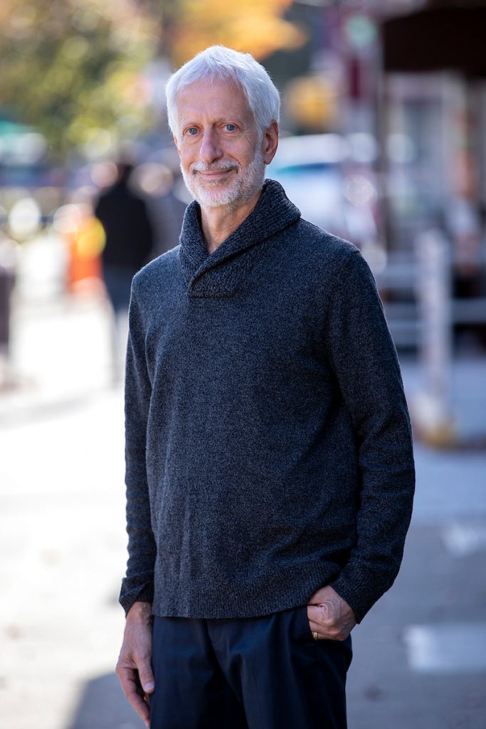 Older man with gray hair and beard standing on a city sidewalk, wearing a dark knit sweater and looking at the camera.
