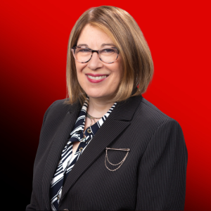 Portrait of a smiling woman with blonde hair and glasses, wearing a dark pinstripe blazer against a red background.