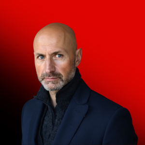 Portrait of a bald man with a short gray beard in a dark blazer, posed against a red-to-black gradient background.