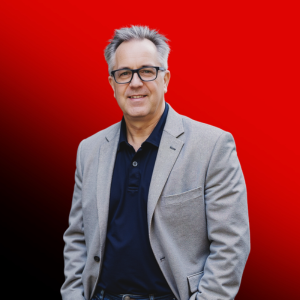 Portrait of a smiling middle-aged man with gray hair and glasses, wearing a navy polo and light gray blazer, against a red gradient background.