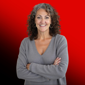 Smiling woman with curly gray hair wearing a gray sweater, arms crossed, standing against a red gradient backdrop.