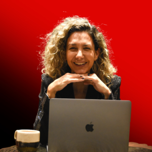 Smiling woman with curly hair sits at a desk with a silver MacBook, hands folded, coffee cup nearby, against a bright red background.