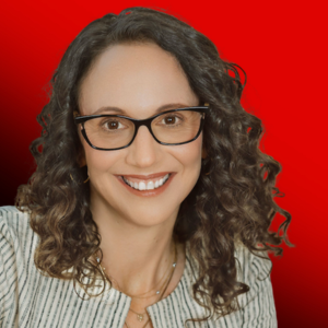 Smiling woman with curly brown hair and glasses, wearing a striped blazer, against a solid red background.