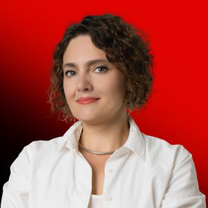 Portrait of a woman with short curly hair in a white shirt, standing against a red background with a confident expression.